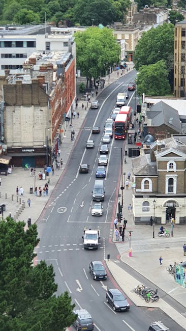 Aerial view of a daytime urban street scene in Sydenham SE26, featuring a mix of classic and modern buildings with brick facades and storefronts along a tree-lined road. The street has multiple lanes 
