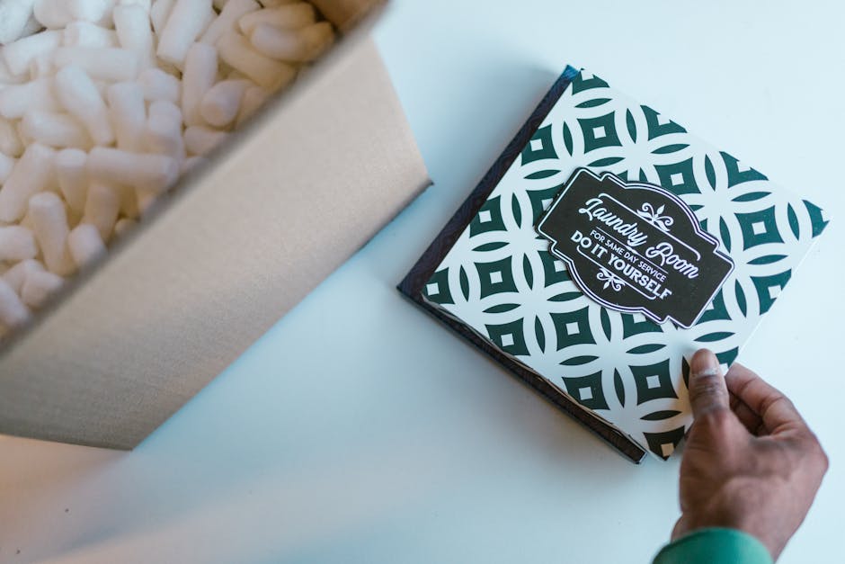 A hand holding a square floral gift box with a black and white geometric pattern and a label that reads 'Laundry Room for Same Day Service Do It Yourself'. The box is positioned on a light-colored sur