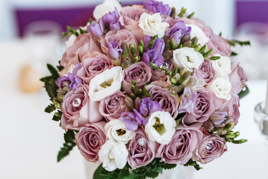 A close-up of a floral bouquet featuring light purple and lavender roses, white lisianthus, and small purple irises, accented with green foliage and decorative crystal embellishments. The arrangement 