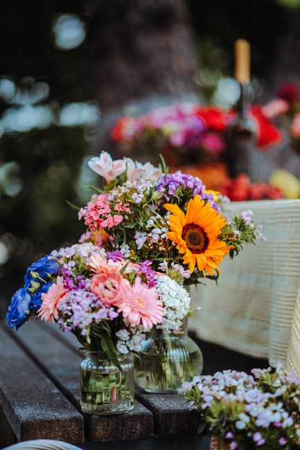 Two glass jars filled with fresh floral bouquets placed on a rustic wooden surface outdoors. The bouquets feature a variety of colorful flowers including a prominent bright yellow sunflower, pink and 