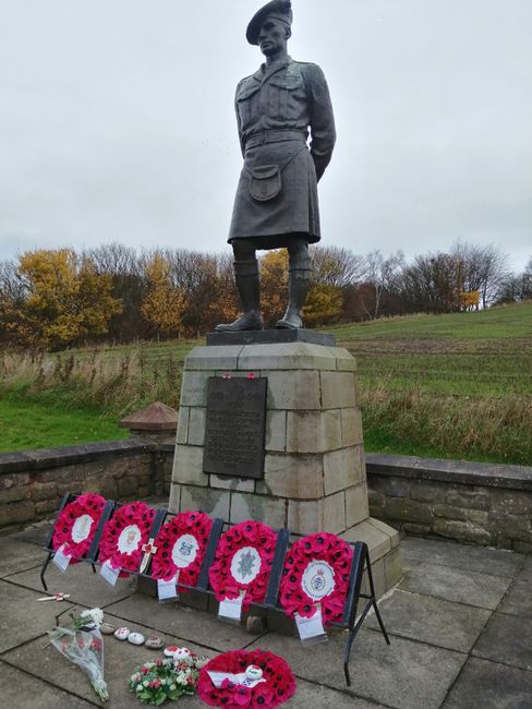 A bronze statue of a woman in military uniform standing on a stone plinth, with a plaque beneath. In front of the monument, there are five arranged floral wreaths predominantly made of bright pink pop